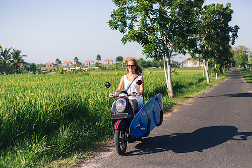 young smiling woman riding scooter with surfing board