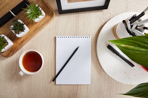 top view of green plants, cup of tea and blank notebook with pencils and pens on wooden surface