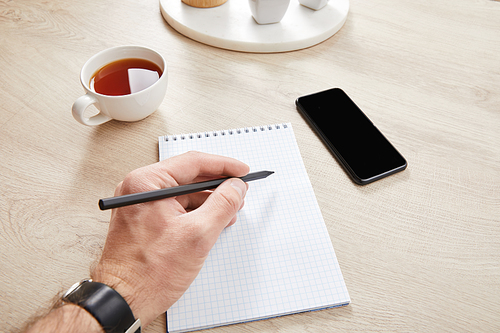 cropped view of man writing in notebook near cup of tea and smartphone on wooden surface