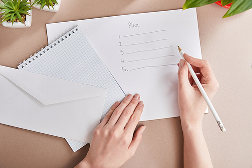 cropped view of woman writing plan on paper near green plants, envelope, blank notebook on beige surface