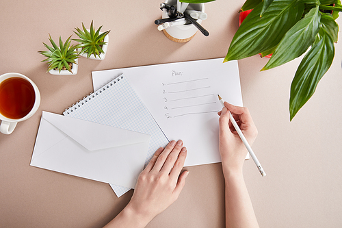 cropped view of woman writing plan on paper near green plants, cup of tea, envelope, blank notebook on beige surface