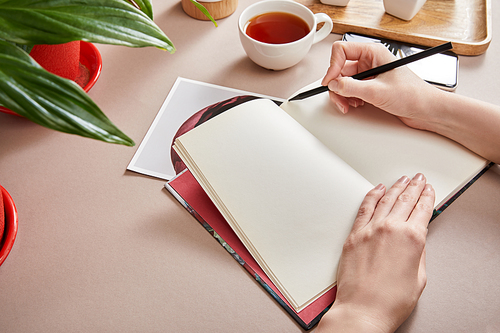cropped view of woman writing in planner near green plant, cup of tea, smartphone on beige surface