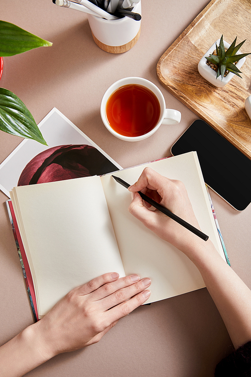 cropped view of woman writing in planner near green plants on wooden board, cup of tea, smartphone on beige surface