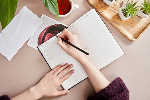 cropped view of woman writing in planner near green plants on wooden board, cup of tea, envelope on beige surface