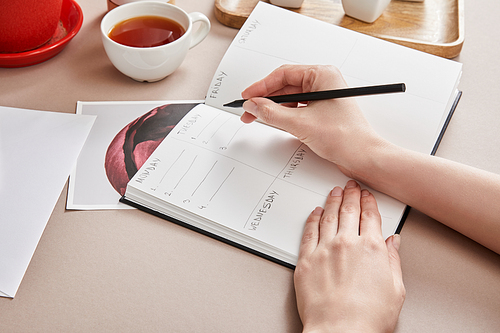 cropped view of woman writing in planner near cup of tea on beige surface