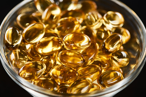 close up view of golden fish oil capsules in glass bowl isolated on black