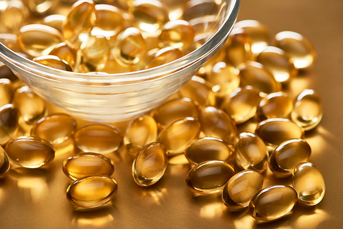 close up view of shiny fish oil capsules scattered from glass bowl on golden background
