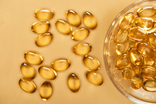 top view of shiny golden fish oil capsules scattered from glass bowl on yellow background