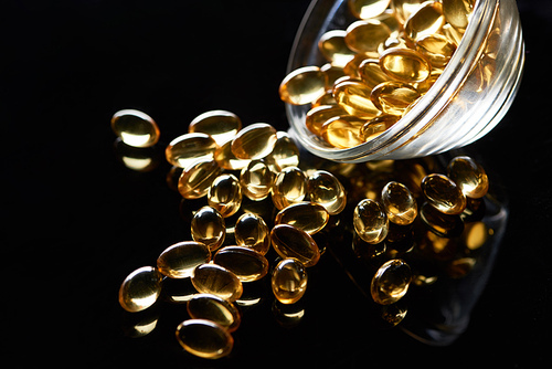 shiny golden fish oil capsules scattered from glass bowl on black background