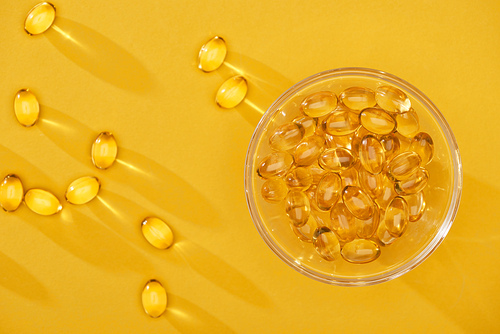 top view of golden shiny fish oil capsules scattered near glass bowl on yellow bright background