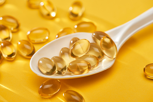 close up view of golden shiny fish oil capsules scattered from spoon on yellow bright background