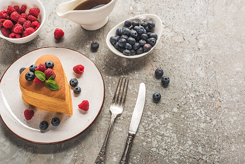 top view of heart shaped pancakes with berries on grey concrete surface with cutlery, maple syrup