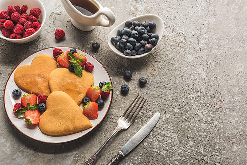 top view of heart shaped pancakes with berries on grey concrete surface with cutlery and maple syrup