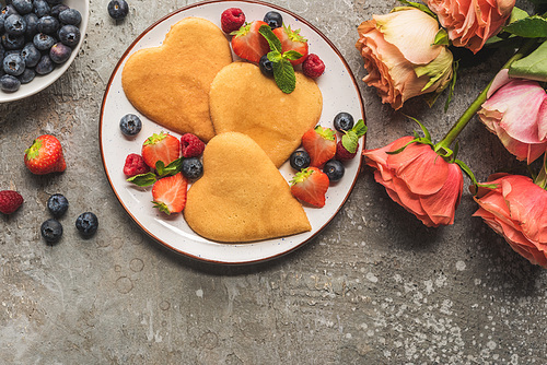 top view of heart shaped pancakes with berries on grey concrete surface near roses