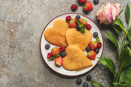 top view of heart shaped pancakes with berries on grey concrete surface near rose