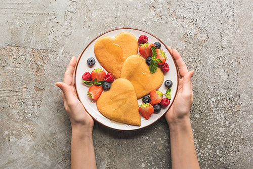 cropped view of woman holding plate with heart shaped pancakes and berries on grey concrete surface