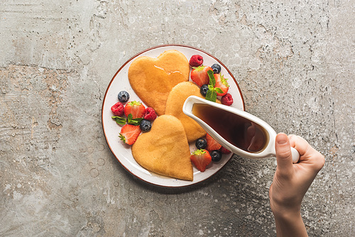 cropped view of woman adding maple syrup to heart shaped pancakes with berries on grey concrete surface