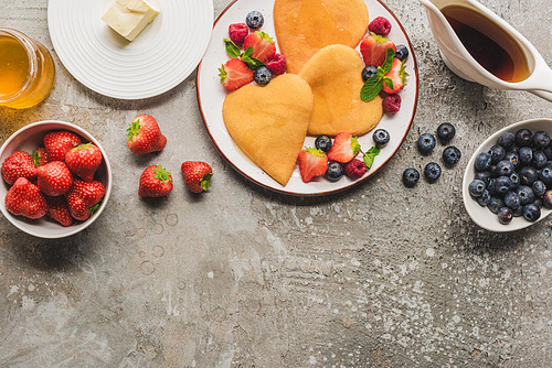 top view of heart shaped pancakes with berries on grey concrete surface with butter, honey and maple syrup
