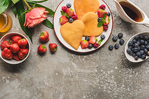 top view of heart shaped pancakes with berries on grey concrete surface with blooming rose, honey and maple syrup