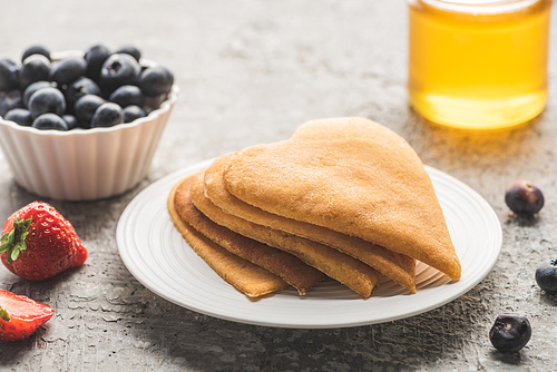 delicious heart shaped pancakes with berries on plate near honey on grey concrete surface