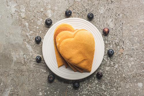 top view of delicious heart shaped pancakes on plate near blueberries on grey concrete surface