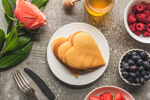 top view of heart shaped pancakes with berries, honey, cutlery and rose on grey concrete surface