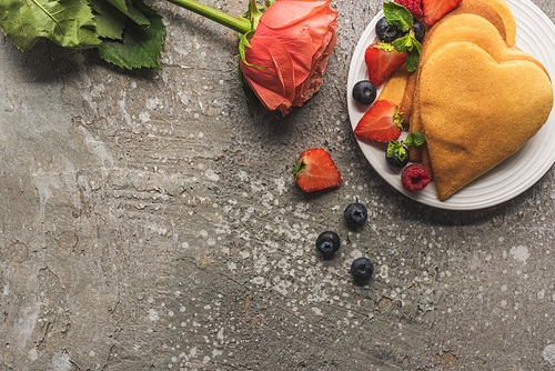 top view of heart shaped pancakes with fresh berries on grey concrete surface with rose