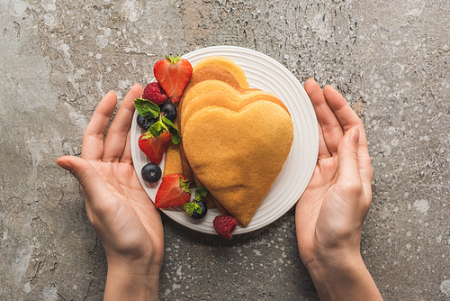 partial view of woman holding plate with heart shaped pancakes and tasty berries on grey concrete surface