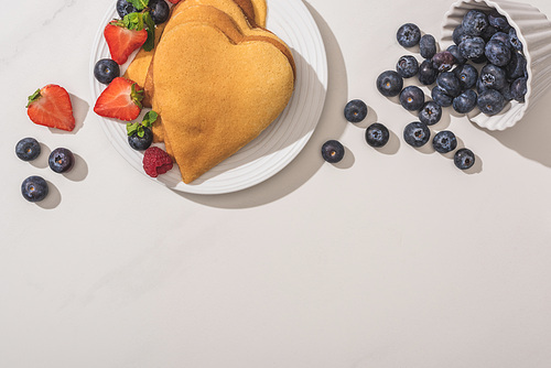 top view of delicious heart shaped pancakes near scattered blueberries on white background