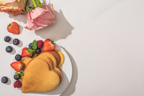 top view of delicious heart shaped pancakes with berries near roses on white background