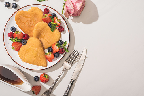 top view of delicious heart shaped pancakes with berries near rose, cutlery and maple syrup on white background