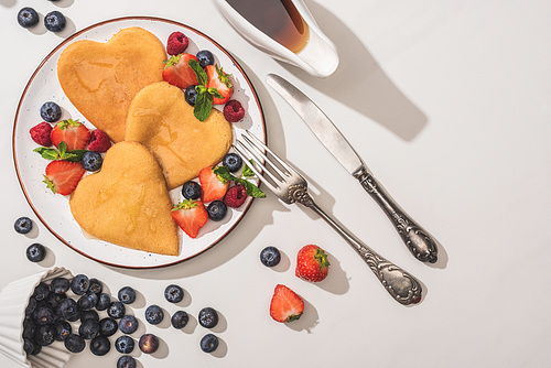 top view of delicious heart shaped pancakes with berries near maple syrup and cutlery on white background