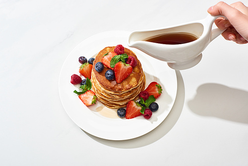 cropped view of woman pouring maple syrup on delicious pancakes with blueberries and strawberries on plate on marble white surface