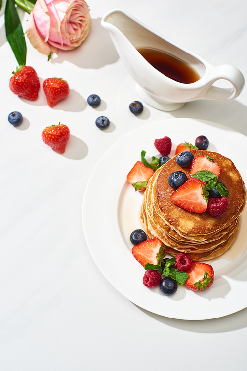 top view of delicious pancakes with maple syrup, blueberries and strawberries on plate near rose flower on marble white surface