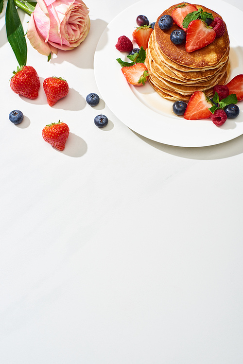 top view of delicious pancakes with blueberries and strawberries on plate near rose flower on marble white surface