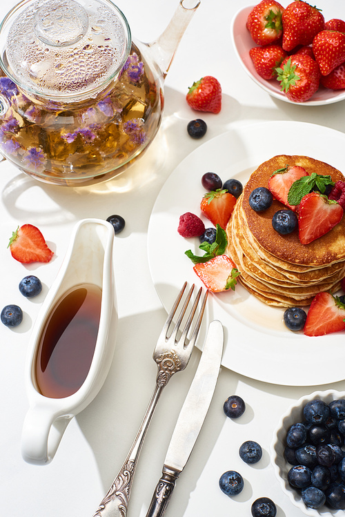 top view of delicious pancakes with maple syrup, blueberries and strawberries on plate near herbal tea in teapot, fork and knife on marble white surface