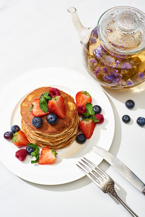 top view of delicious pancakes with maple syrup, blueberries and strawberries on plate near herbal tea in teapot, fork and knife on marble white surface