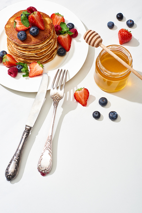 top view of delicious pancakes with honey, blueberries and strawberries on plate near fork and knife on marble white surface