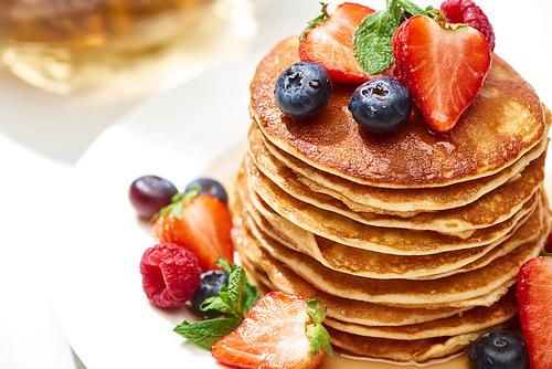 close up view of delicious pancakes with honey, blueberries and strawberries on plate on white surface