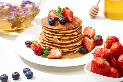 selective focus of delicious pancakes with honey, blueberries and strawberries on plate on white surface