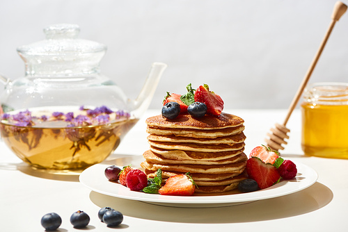 selective focus of delicious pancakes with honey, blueberries and strawberries on plate near herbal tea on white surface isolated on grey