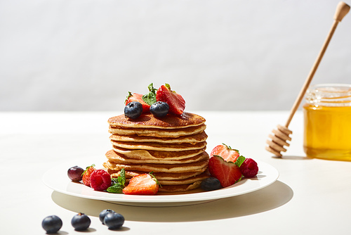 selective focus of delicious pancakes with blueberries and strawberries on plate near honey on white surface isolated on grey