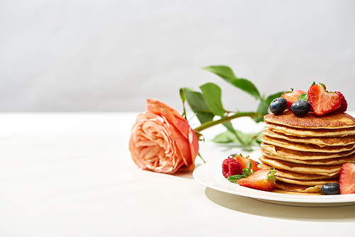 selective focus of delicious pancakes with blueberries and strawberries on plate near rose on white surface isolated on grey