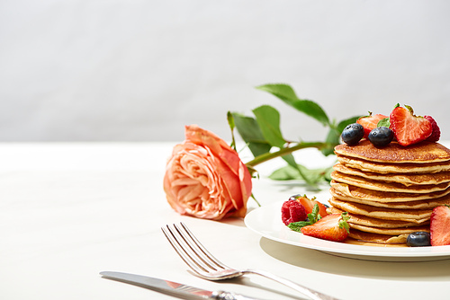 selective focus of delicious pancakes with blueberries and strawberries on plate near rose flower and cutlery on white surface isolated on grey
