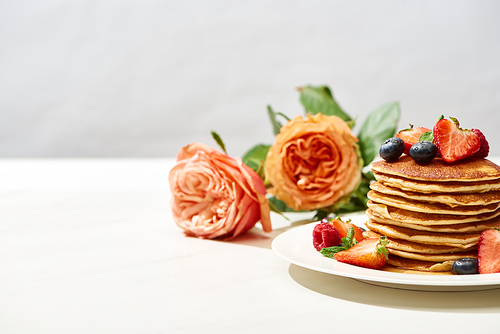 selective focus of delicious pancakes with blueberries and strawberries on plate near rose flowers on white surface isolated on grey