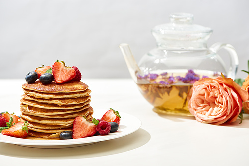 selective focus of delicious pancakes with blueberries and strawberries on plate near rose flowers and herbal tea on white surface isolated on grey