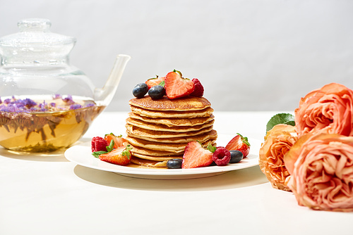 selective focus of delicious pancakes with blueberries and strawberries on plate near rose flowers and herbal tea on white surface isolated on grey