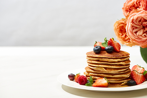 delicious pancakes with blueberries and strawberries on plate near rose flowers on white surface isolated on grey