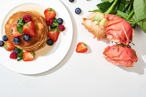 top view of delicious pancakes with honey, blueberries and strawberries on plate near roses on marble white surface