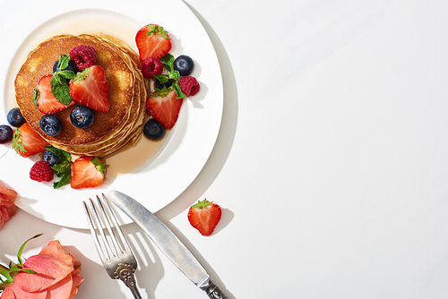 top view of delicious pancakes with honey, blueberries and strawberries on plate near cutlery and rose on marble white surface